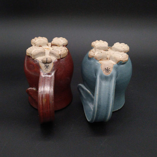 A red and a blue Cat Butt Mug sitting upside down with the feet in the air.