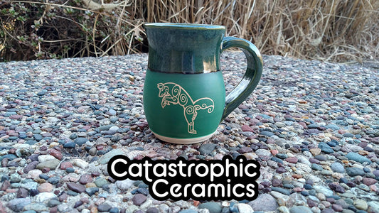 A handmade green mug featuring a Celtic-inspired leaping fox design on a gravel ground with grass in the background.