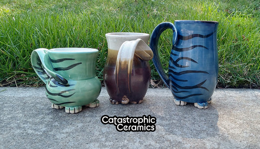 A green striped, a red and cream, and a blue striped cat butt mugs sitting on concrete with grass in the background.